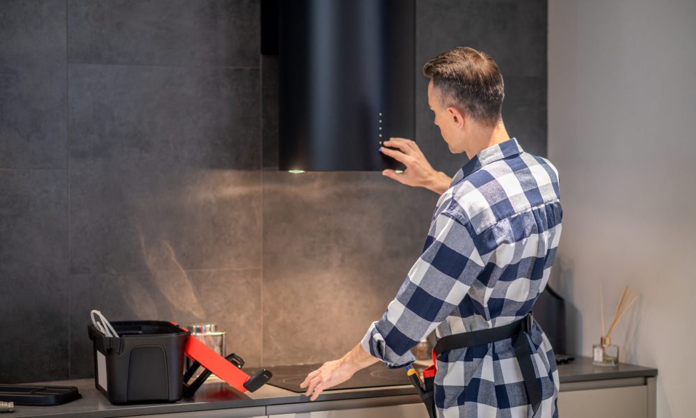 Control panel. Back view of man in plaid shirt touching control panel of black kitchen hood hanging on wall in house